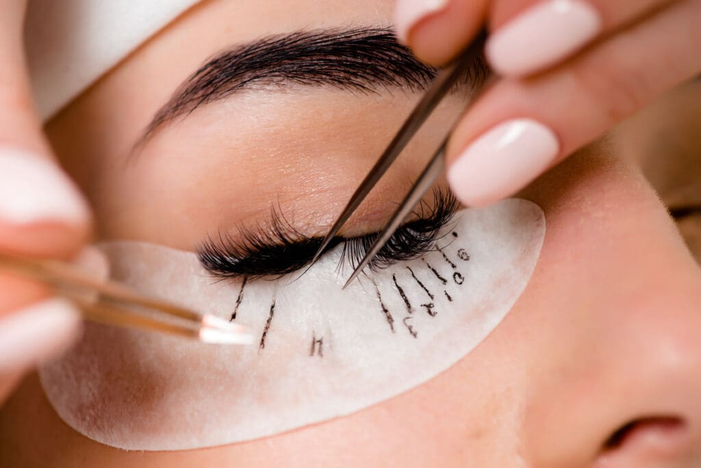 Close-up of a person receiving eyelash extensions; tweezers are used to apply individual lashes while a numbered pad protects the skin under the closed eye.
