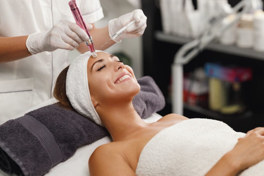 A woman lies smiling on a spa bed with a towel wrapped around her body and a headband on, while a cosmetologist wearing gloves performs a facial treatment using a microneedling device on her forehead.