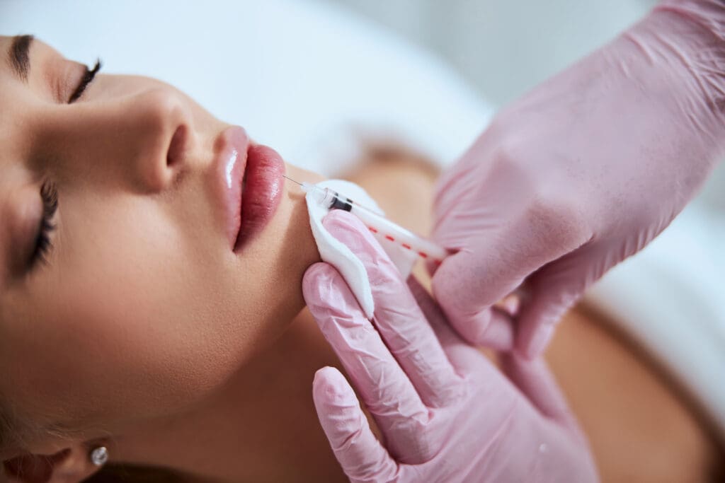 A close-up of a woman receiving a cosmetic lip injection. She has her eyes closed while a person wearing pink gloves holds her chin and injects her lips with a syringe, using a white cloth for support.
