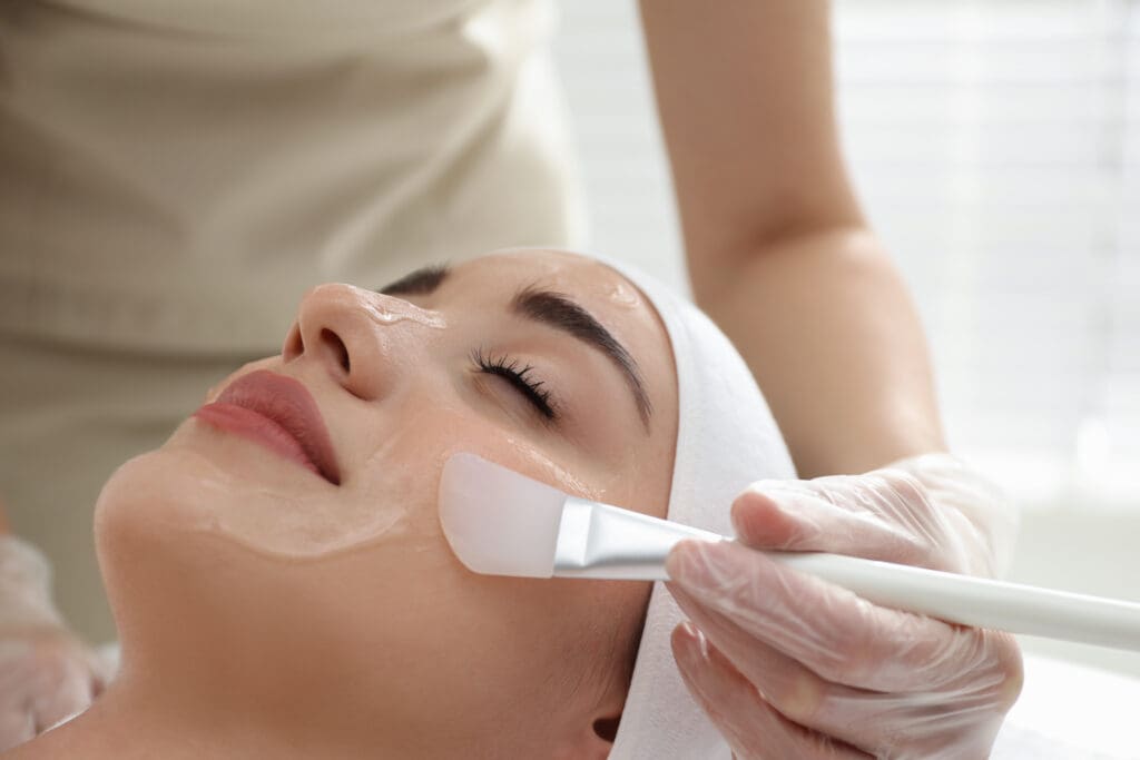 A person with closed eyes is lying down while a gloved hand applies a facial mask with a brush to their cheek. The person is wearing a white headband.