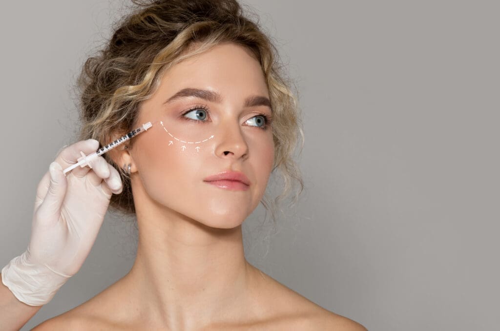 A woman with curly hair receives a cosmetic injection near her eye. White arrows are drawn under her eye, and a gloved hand holds the syringe. She looks slightly to the side against a plain gray background.