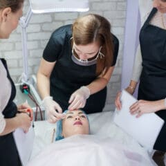 Three women in uniforms observe as a professional performs a facial treatment on a woman lying on a bed, wearing a headwrap, in a bright, clinical setting.