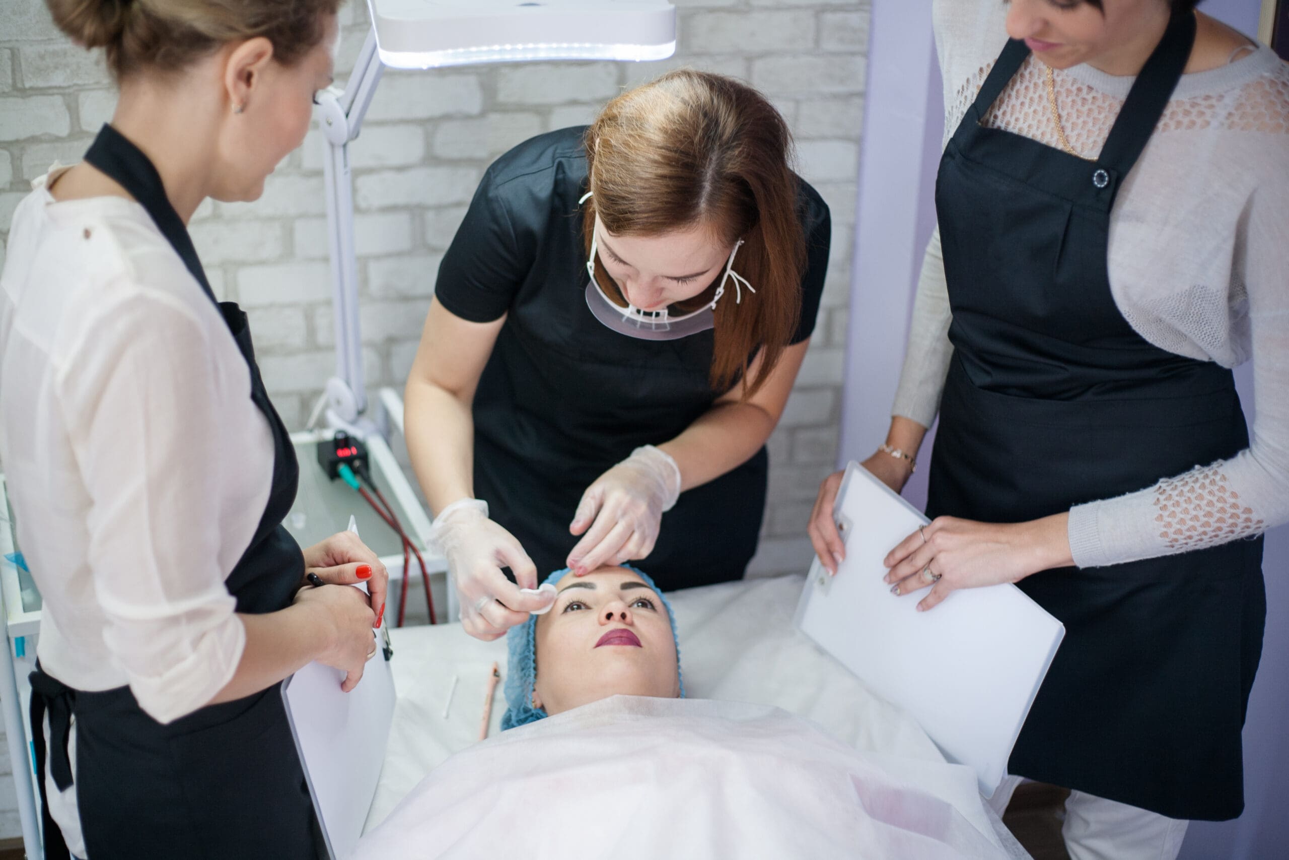 Three women in uniforms observe as a professional performs a facial treatment on a woman lying on a bed, wearing a headwrap, in a bright, clinical setting.