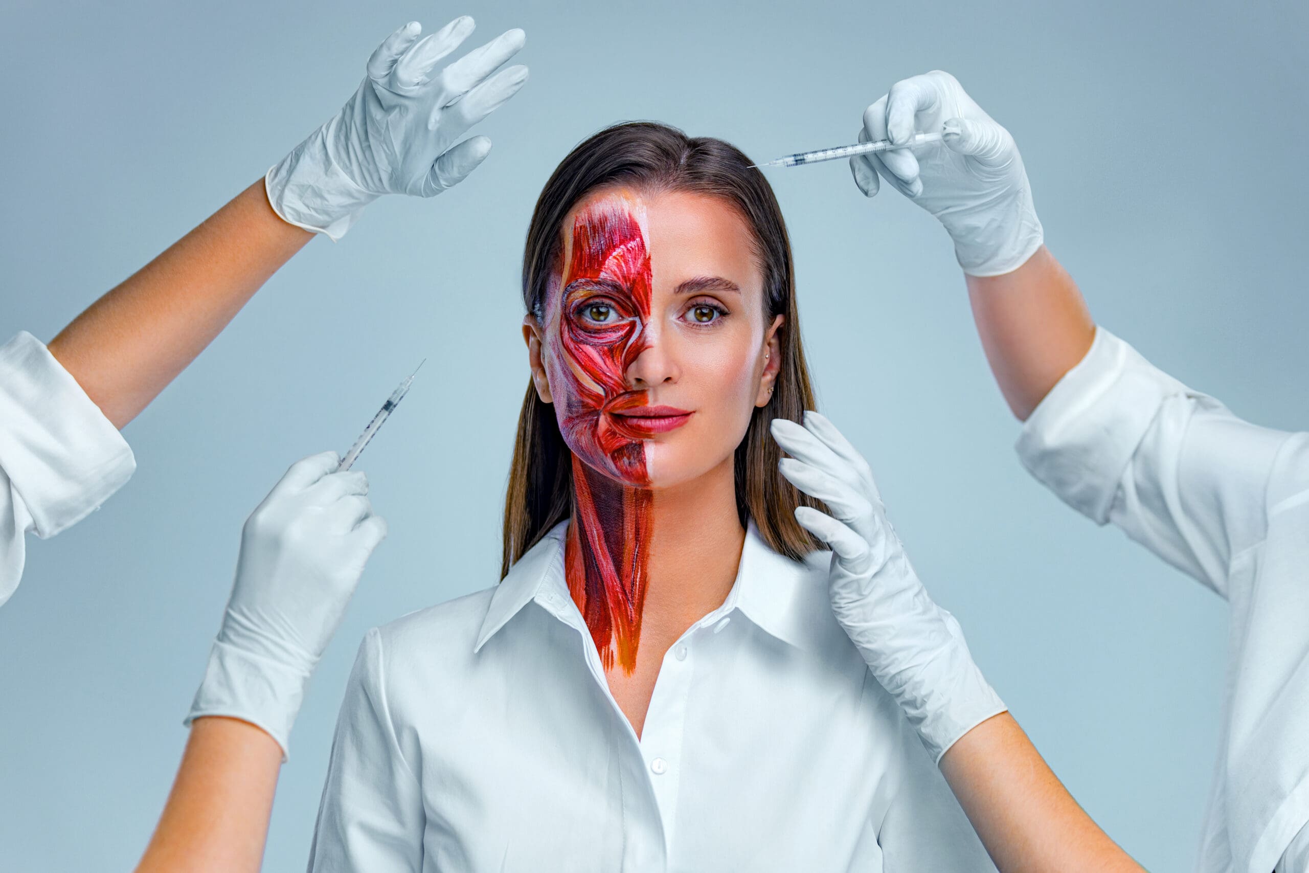 A woman in a white shirt faces the camera as three gloved hands hold syringes near her face. Half of her face is painted to show underlying muscles, highlighting anatomy and cosmetic procedures.