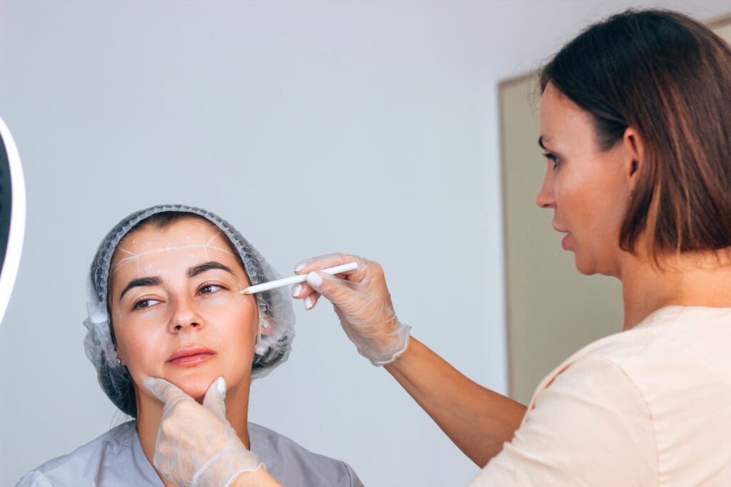 A medical professional wearing gloves marks lines on a woman’s face with a white pencil, preparing her for a cosmetic procedure. The woman wears a hair net and looks slightly to the side.