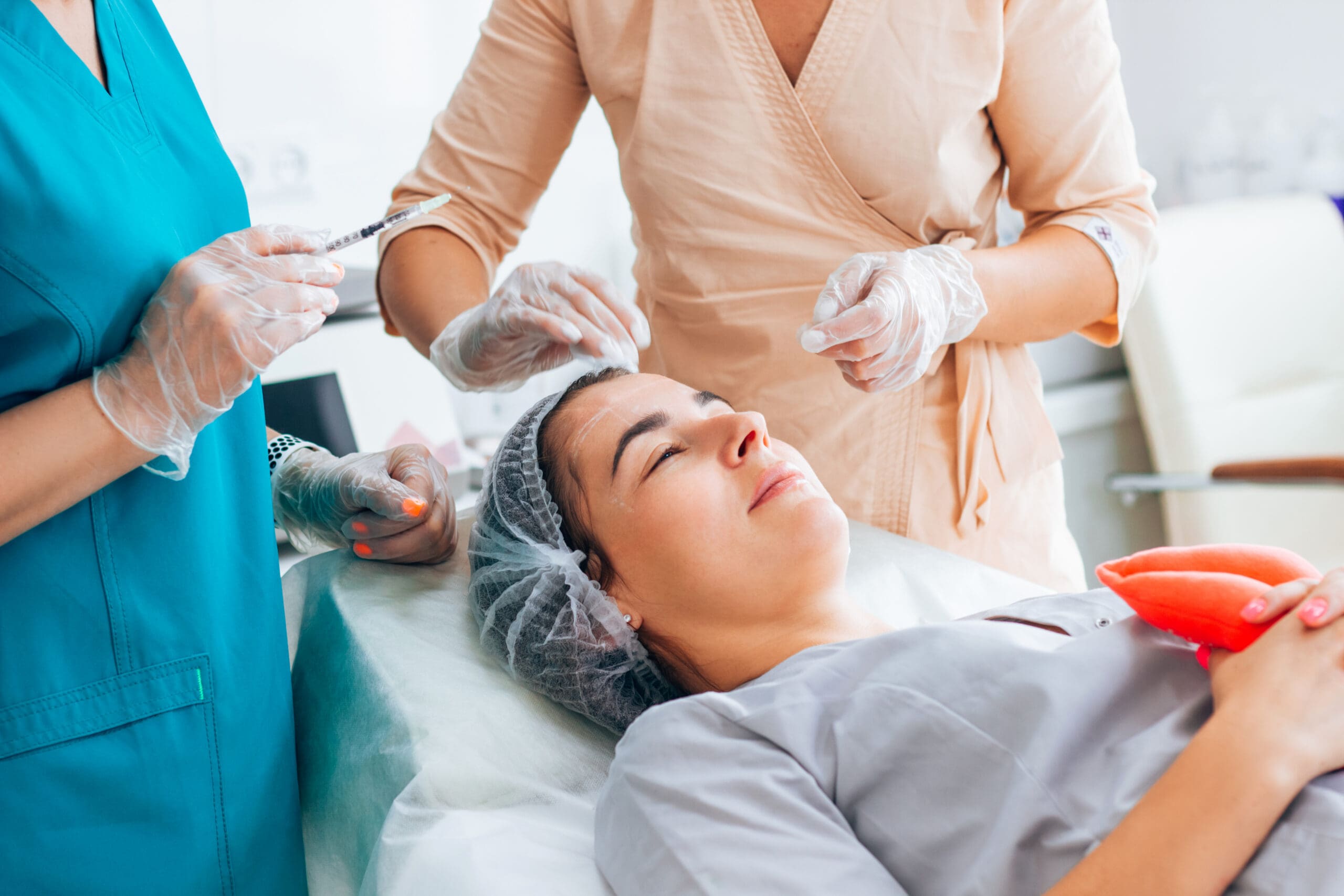 A woman lies on a medical chair with a hair cover, while two healthcare professionals in gloves prepare to give her an injection in her face, suggesting a cosmetic or dermatological procedure.