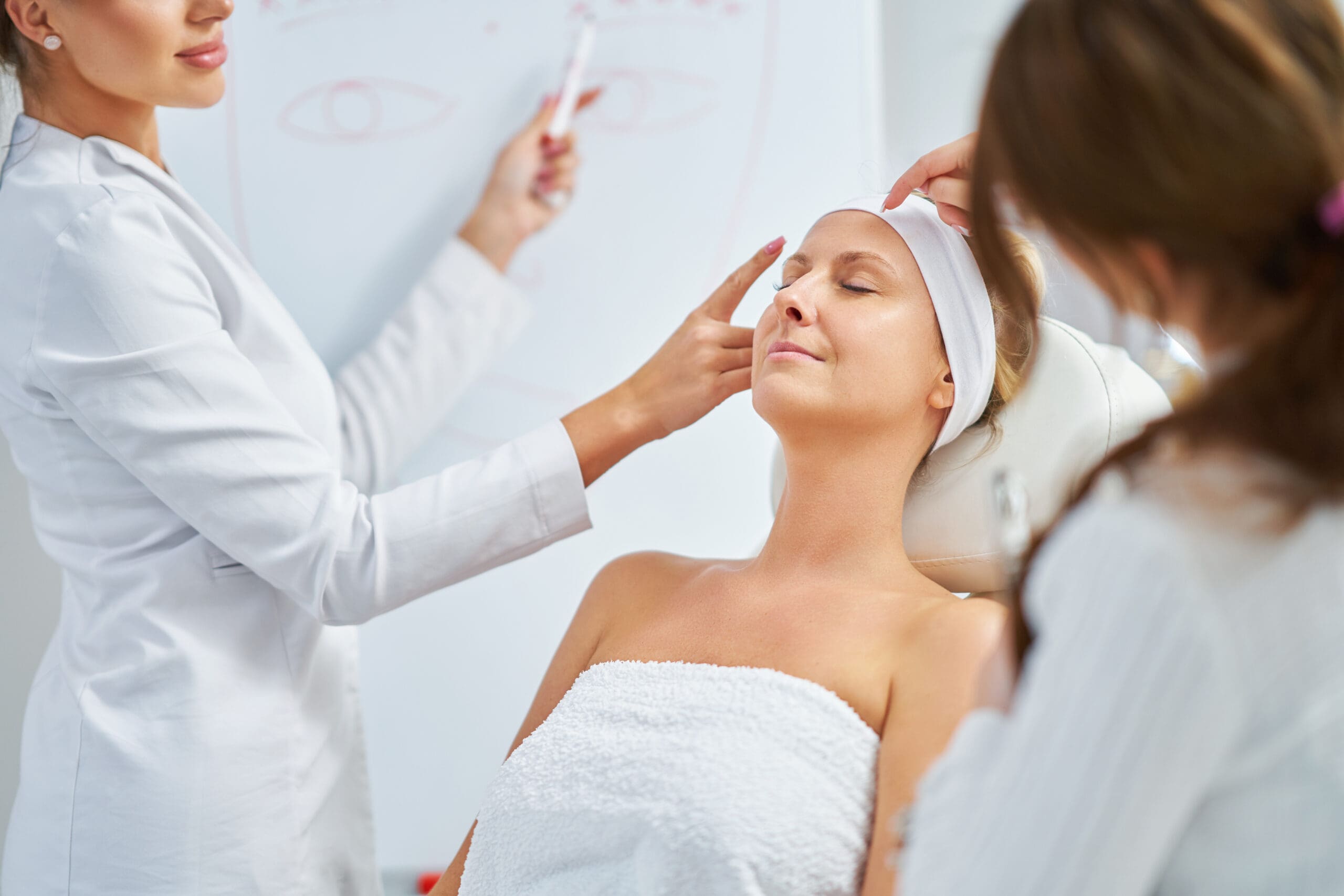 A woman with a headband relaxes in a clinic chair while two professionals in white coats prepare for a facial procedure; a diagram of eyes is visible on a whiteboard in the background.