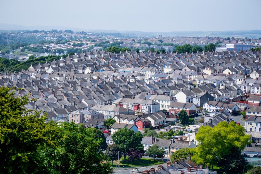 A densely packed neighborhood with rows of grey-roofed houses stretches across the landscape, interspersed with green trees and backed by distant hills under a hazy sky.